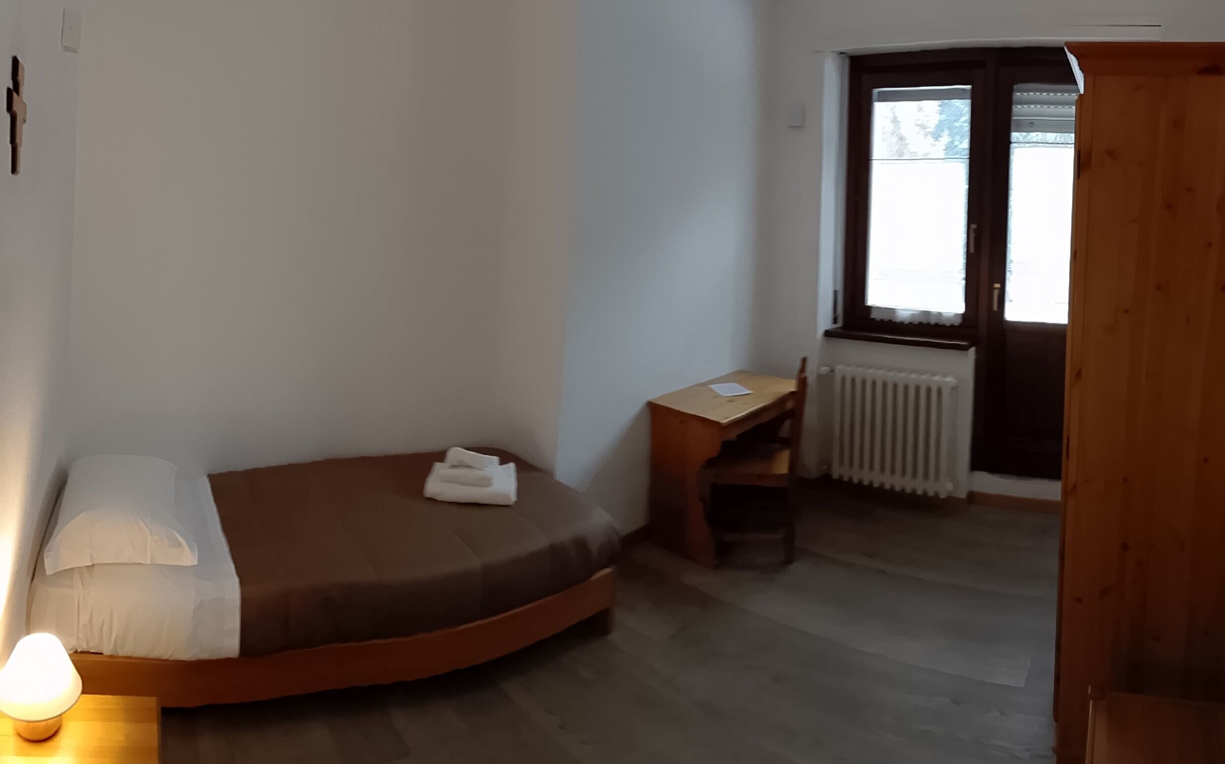Pequeña habitación con cama individual, cabecero de madera, mesita de noche, una silla, ventana con vistas y un radiador.