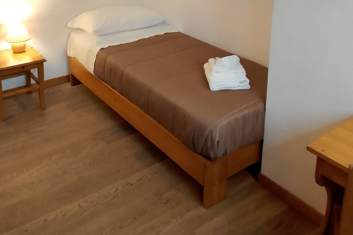 A single bed with a brown mattress and white pillows, a wooden nightstand with a lamp, and a folded towel on the bed.