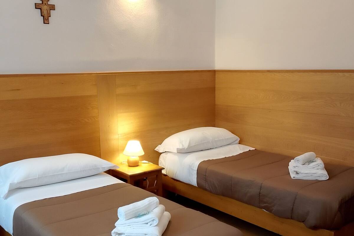 A hotel room with two single beds, a wooden headboard, a wall-mounted cross, and a nightstand with a lamp.