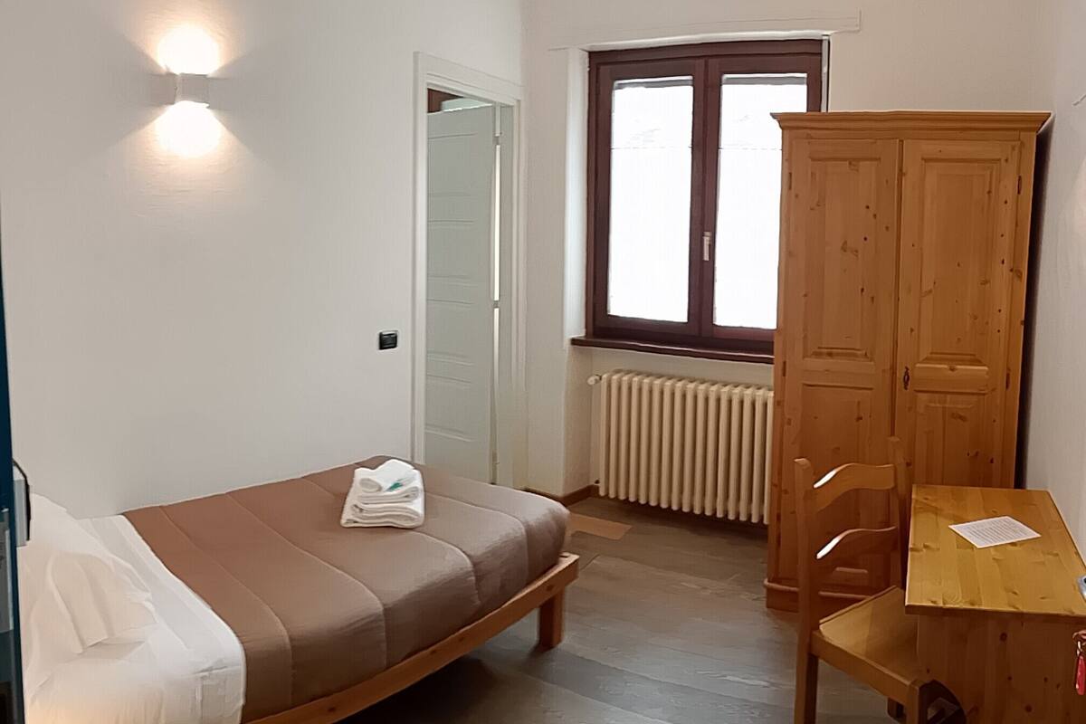 A bedroom with a bed, a wooden wardrobe, a desk, a chair, a radiator, and a window.