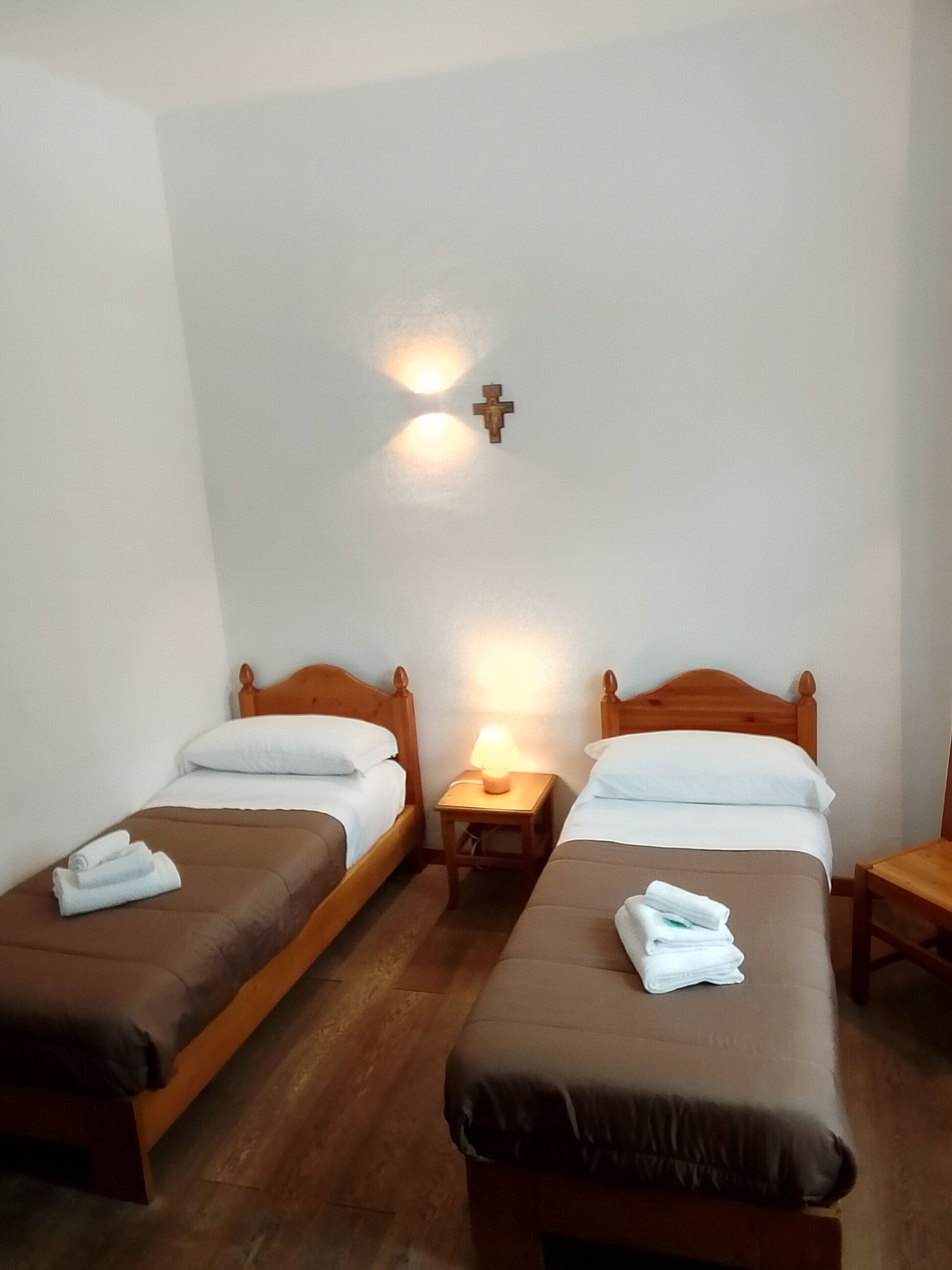 A hotel room with two wooden beds, a nightstand with a lamp, and a wall-mounted cross.