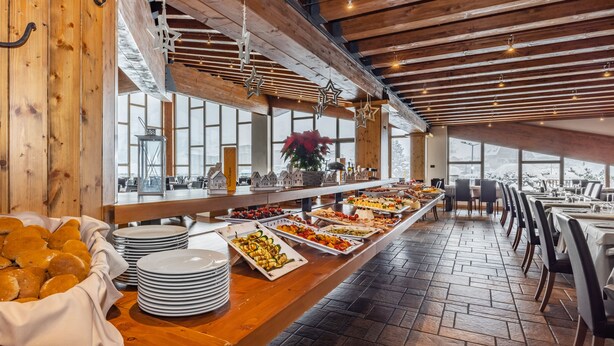 A buffet setup with various food items, a wooden counter, and a view of the snowy landscape through large windows. - Hotel Piandineve (Ponte di Legno)