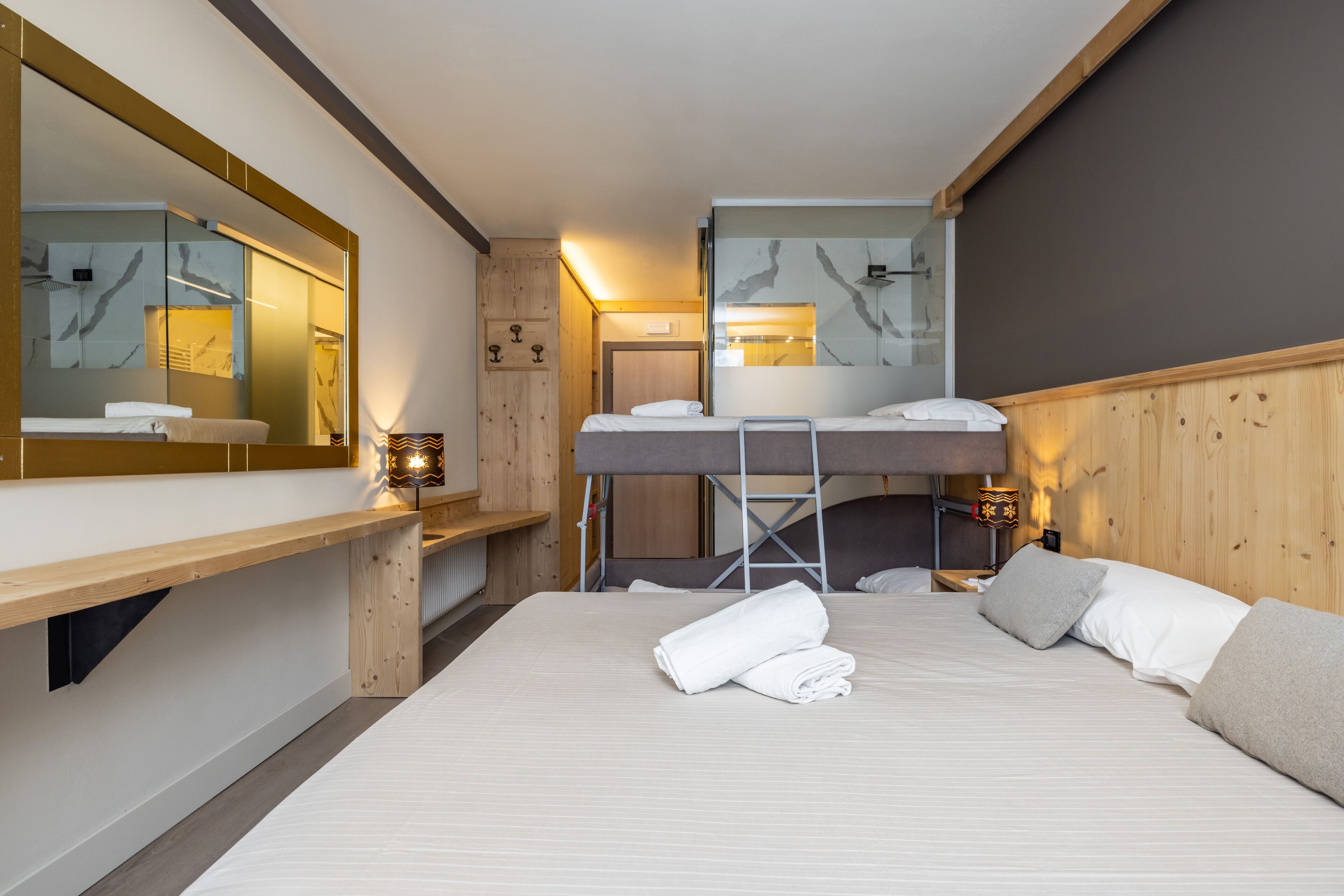 A modern hotel room with a bunk bed, a large mirror, a wooden bench, and a bedside table with a lamp.