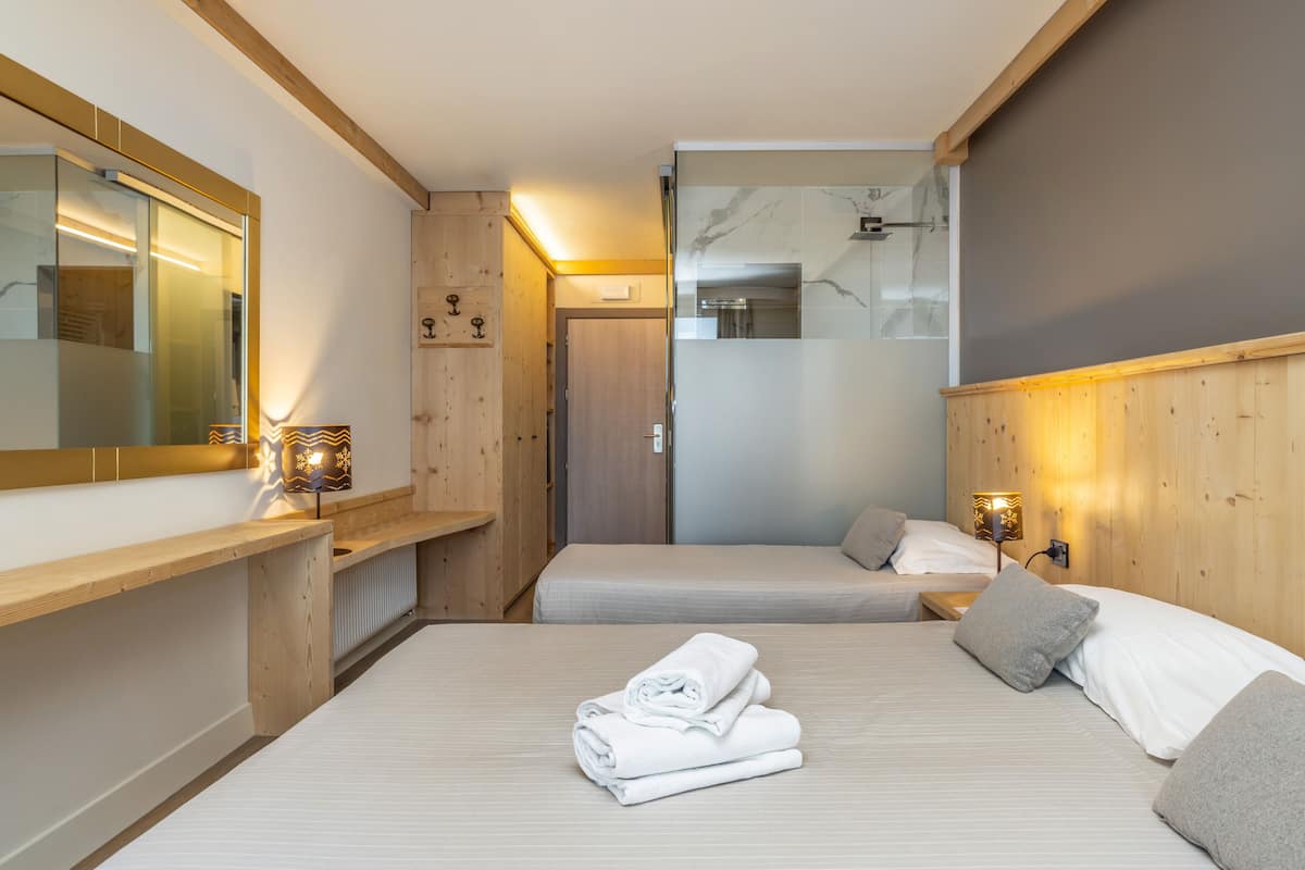 A hotel room with two beds, a wooden bench, a mirror, and a bathroom visible through a frosted glass door.
