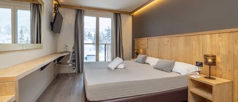 A modern bedroom with a large bed, wooden headboard, a bedside table with a lamp, a TV mounted on the wall, and a window with curtains.