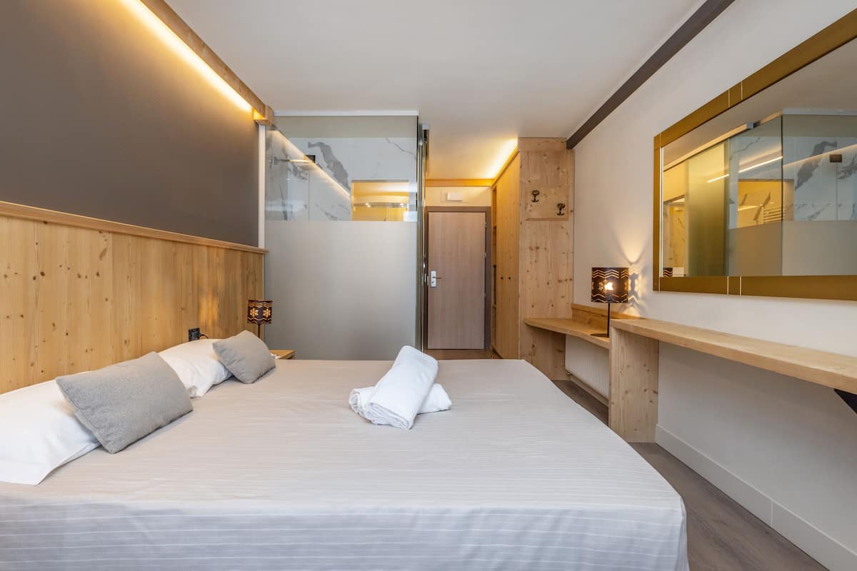 A modern hotel room with a large bed, wooden headboard, a mirror, and a small desk with a lamp.