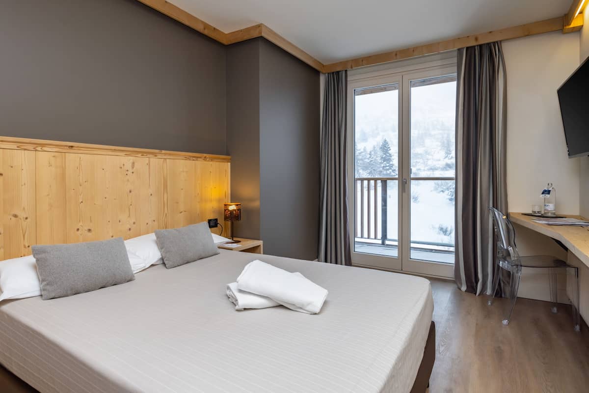A hotel room with a large bed, wooden headboard, a desk with a chair, a TV, and a window with curtains.