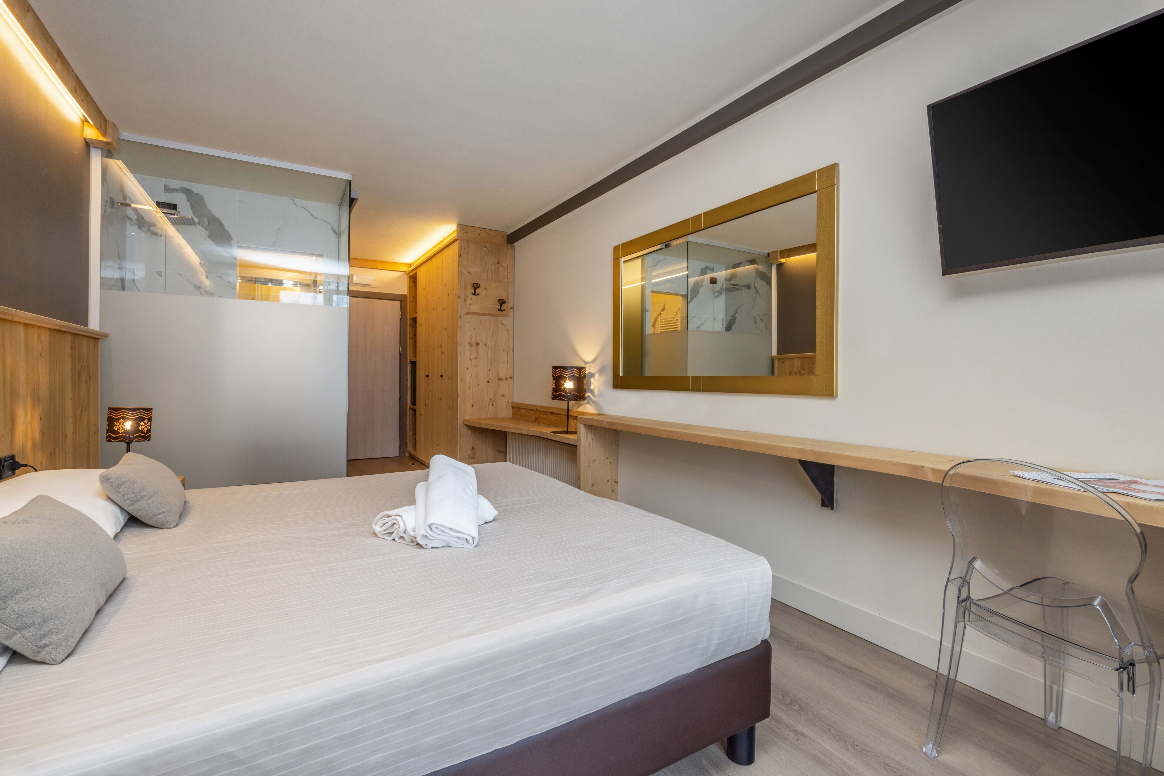 A modern hotel room with a large bed, a wooden desk, a chair, a TV mounted on the wall, and a mirror above the desk.