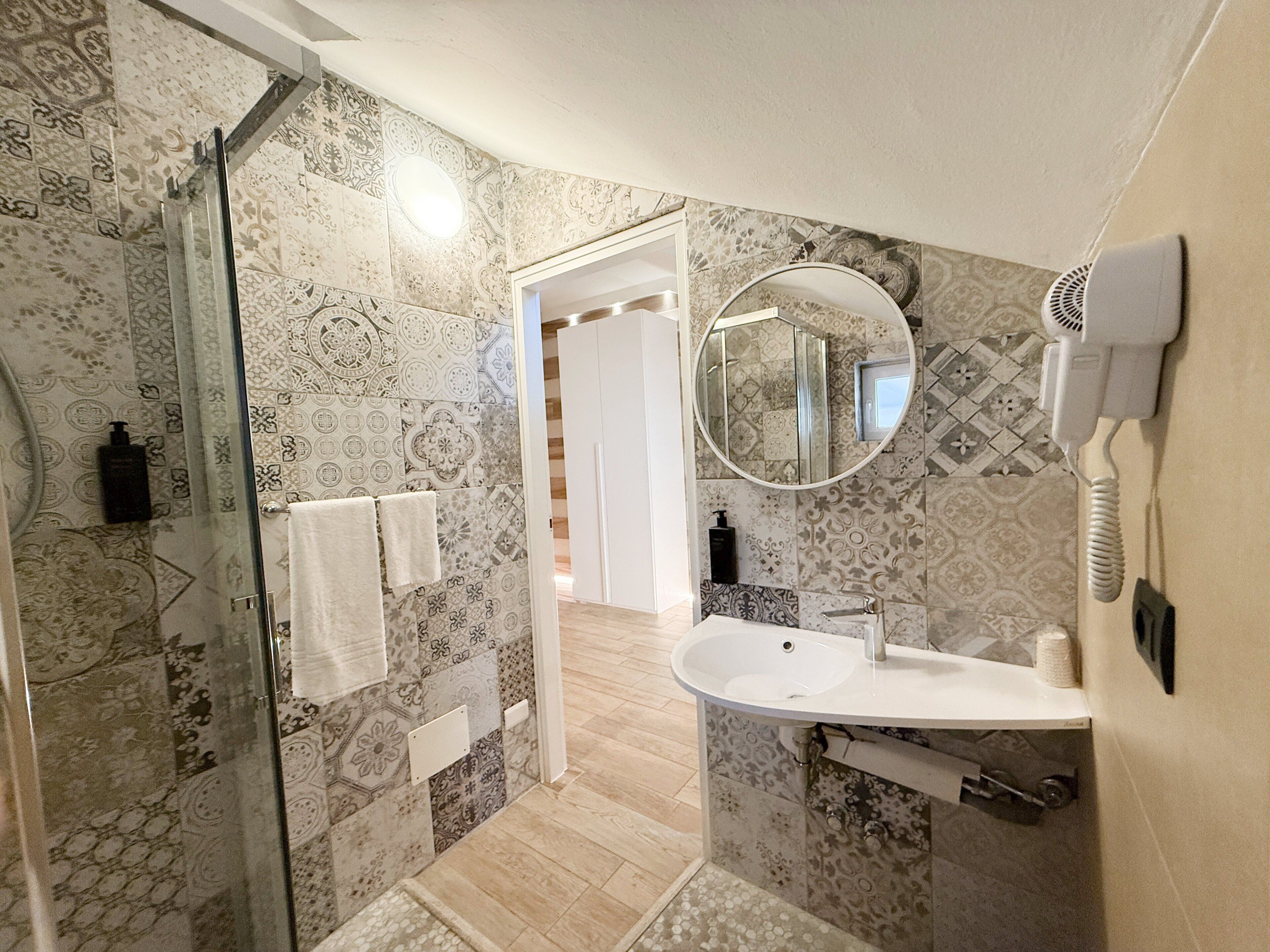A modern bathroom with patterned tiles, a glass shower enclosure, a sink with a mirror, and a hairdryer mounted on the wall.