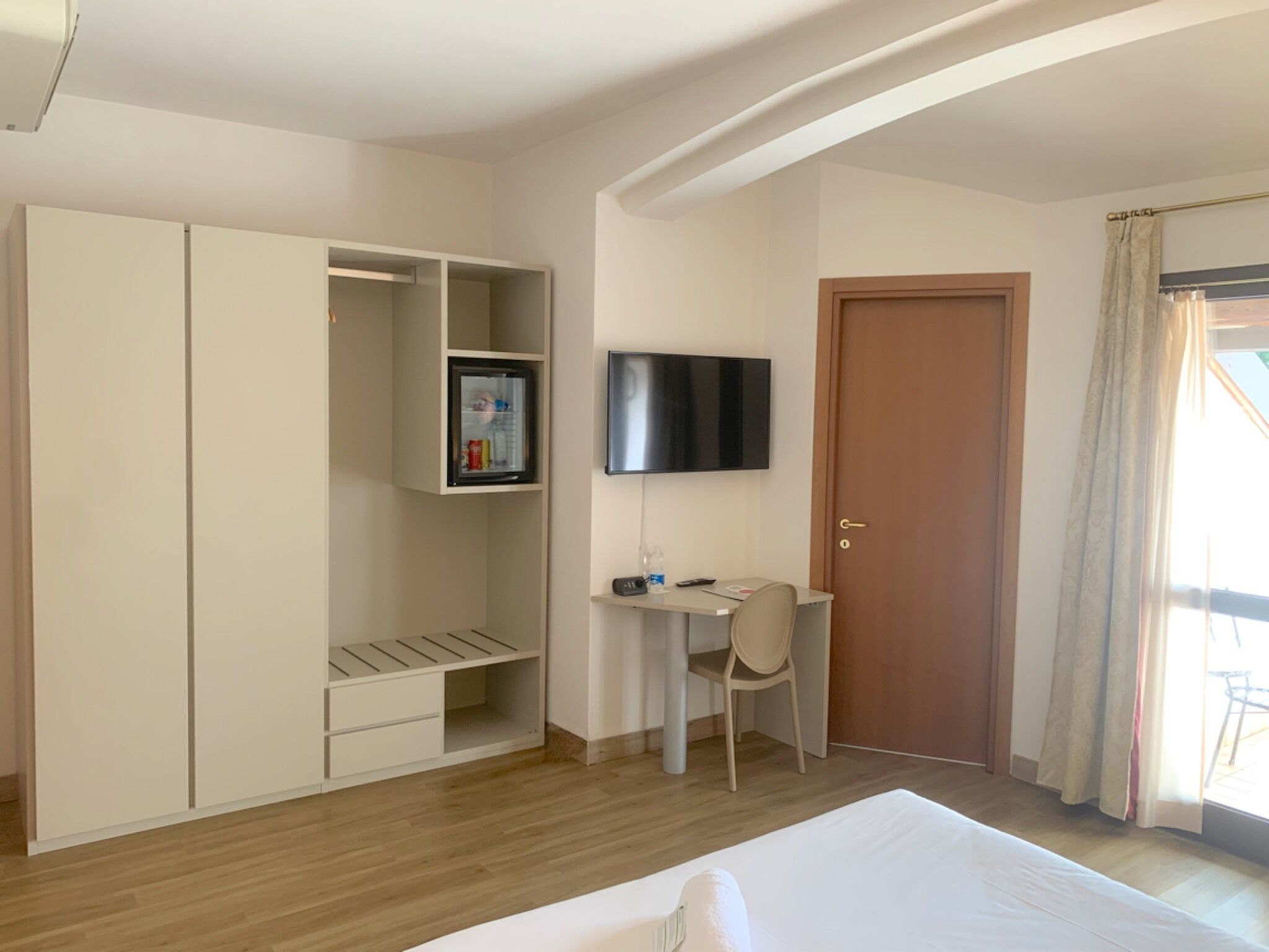 A hotel room with a bed, a desk with a chair, a wardrobe, a television mounted on the wall, and a door.