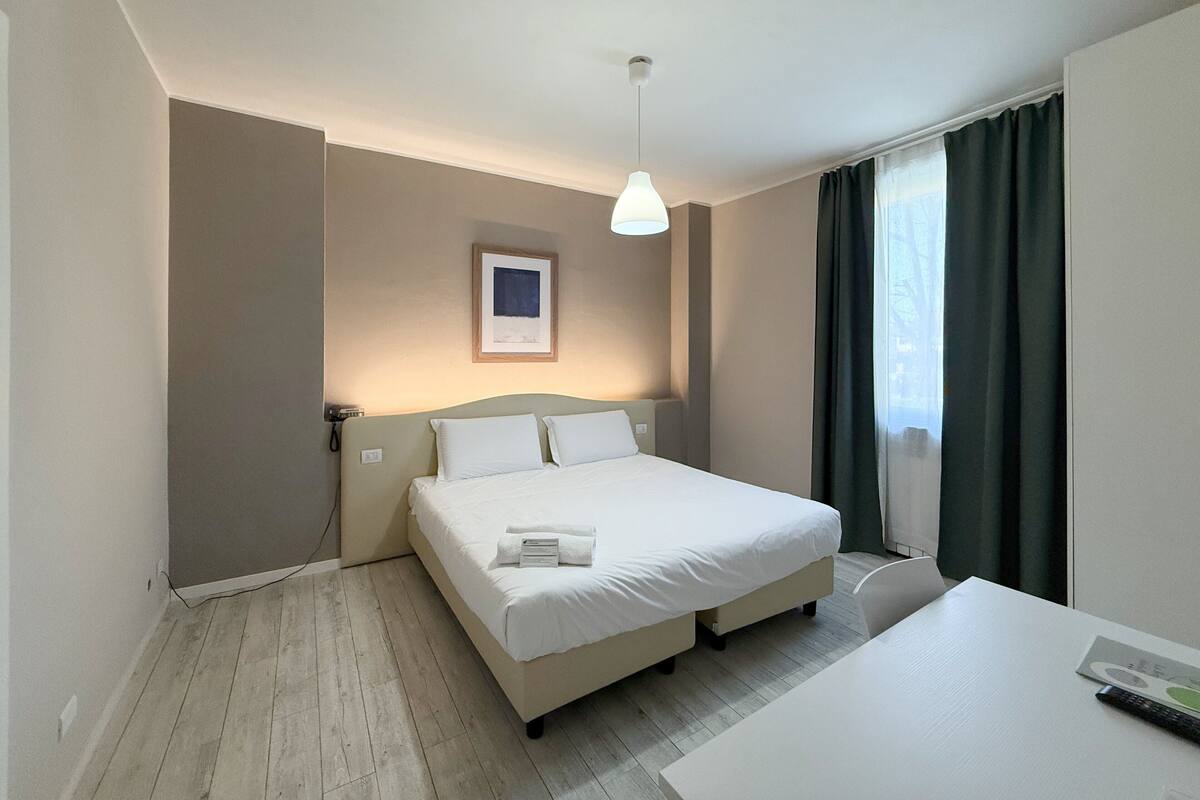 A hotel room with a bed, a desk, a chair, a framed picture on the wall, and a window with curtains.