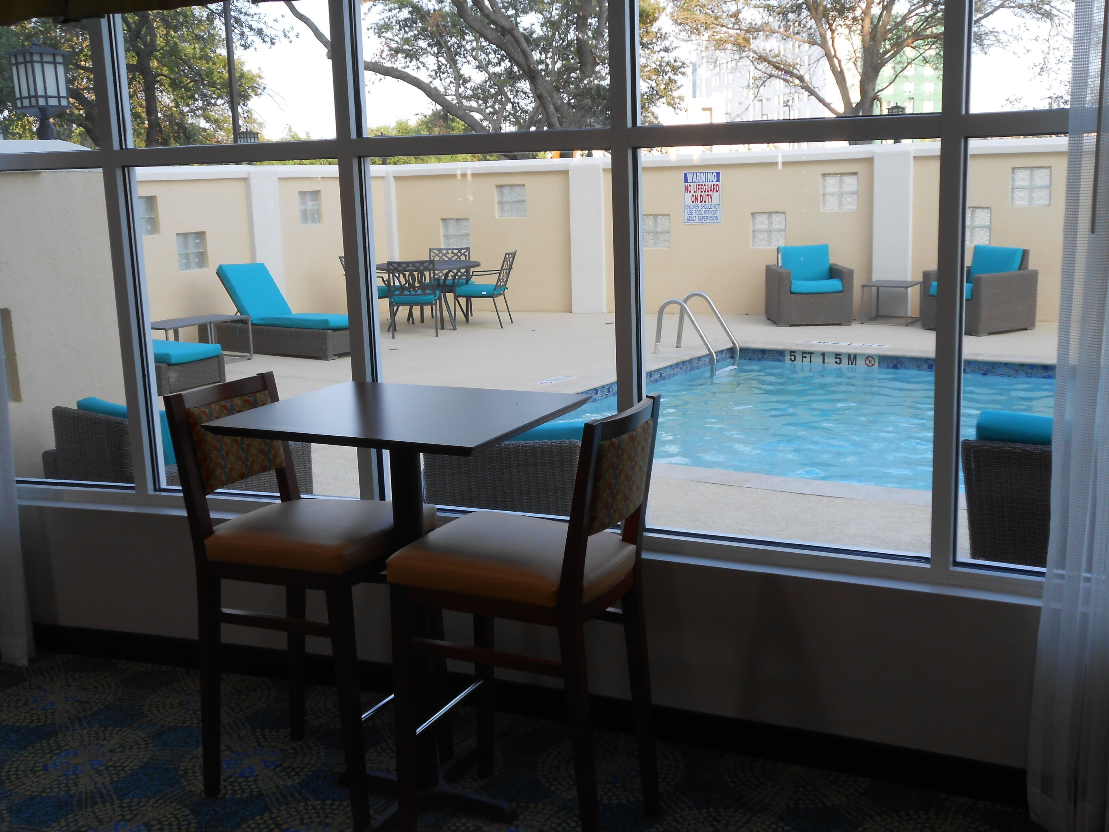 Uma área de piscina com espreguiçadeiras e uma mesa, uma placa de aviso e vista para a piscina através de amplas janelas.