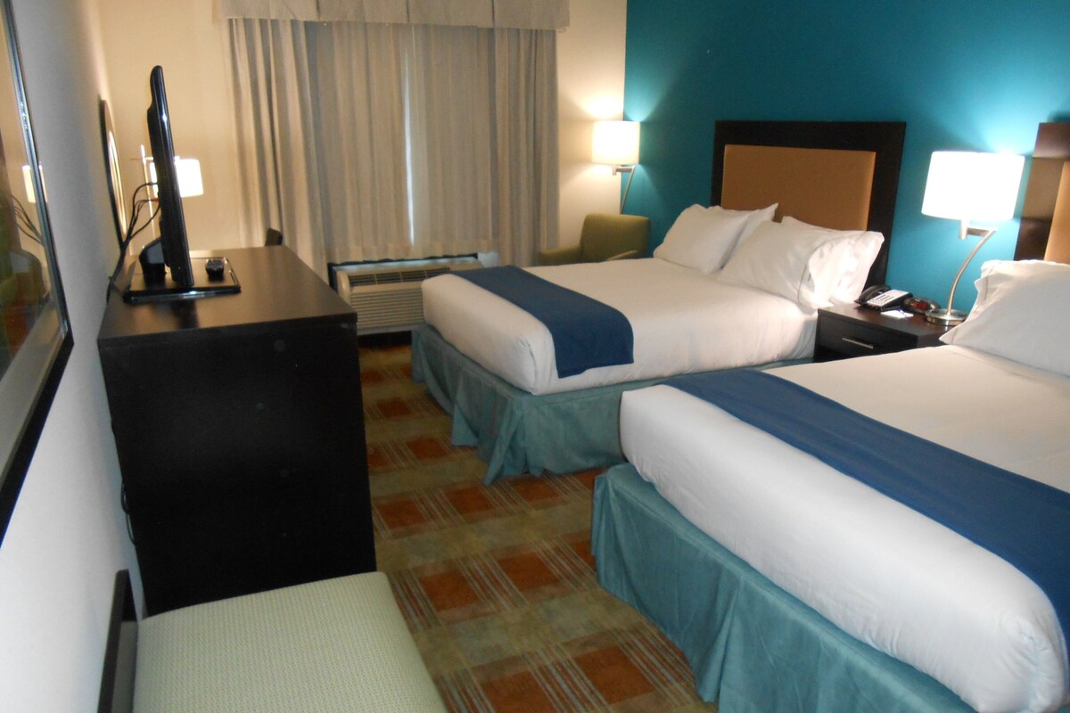 A hotel room with two beds, a desk, a chair, a TV, and a lamp.