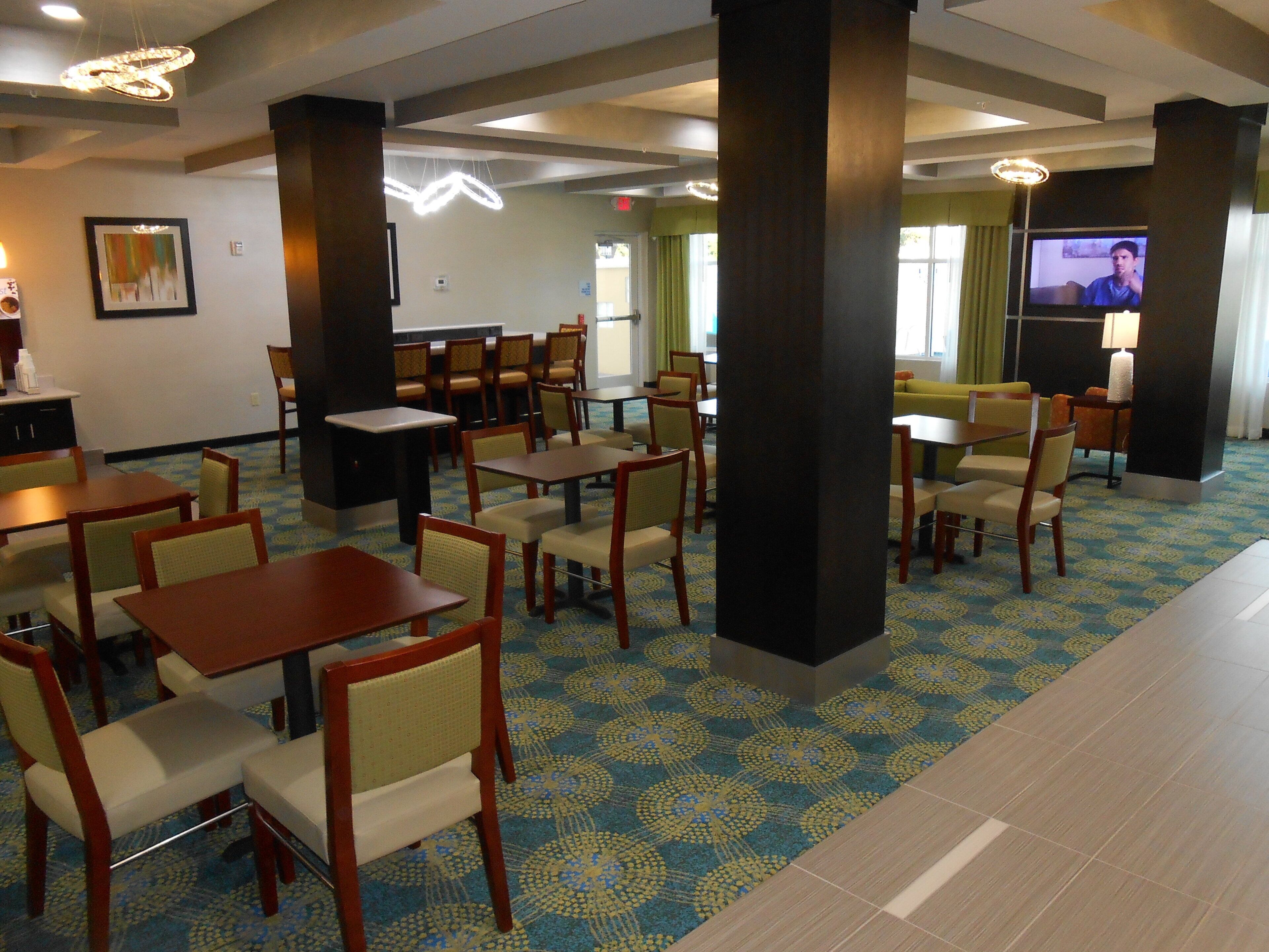 A hotel dining area with tables and chairs, a television screen, and a patterned carpet.