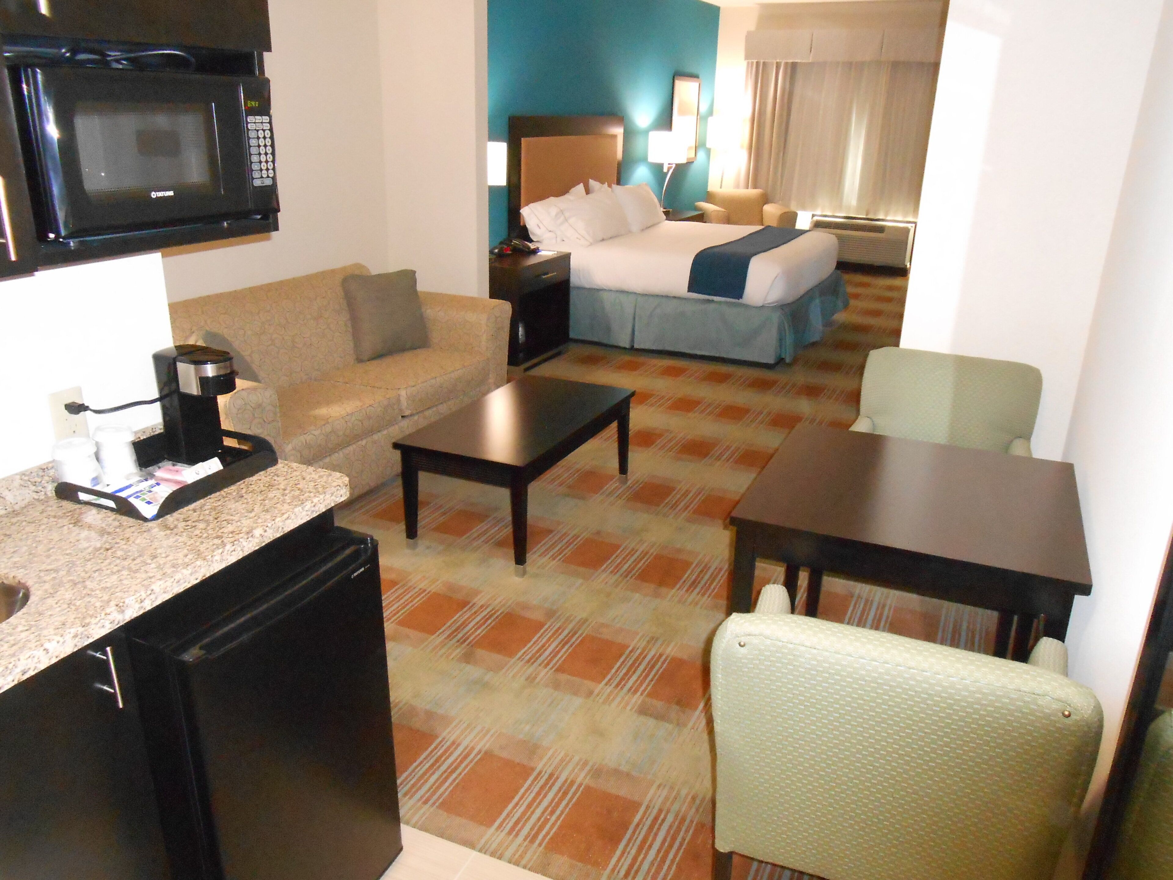 A hotel room with a kitchenette, a bed, a sofa, a dining table, and a chair.
