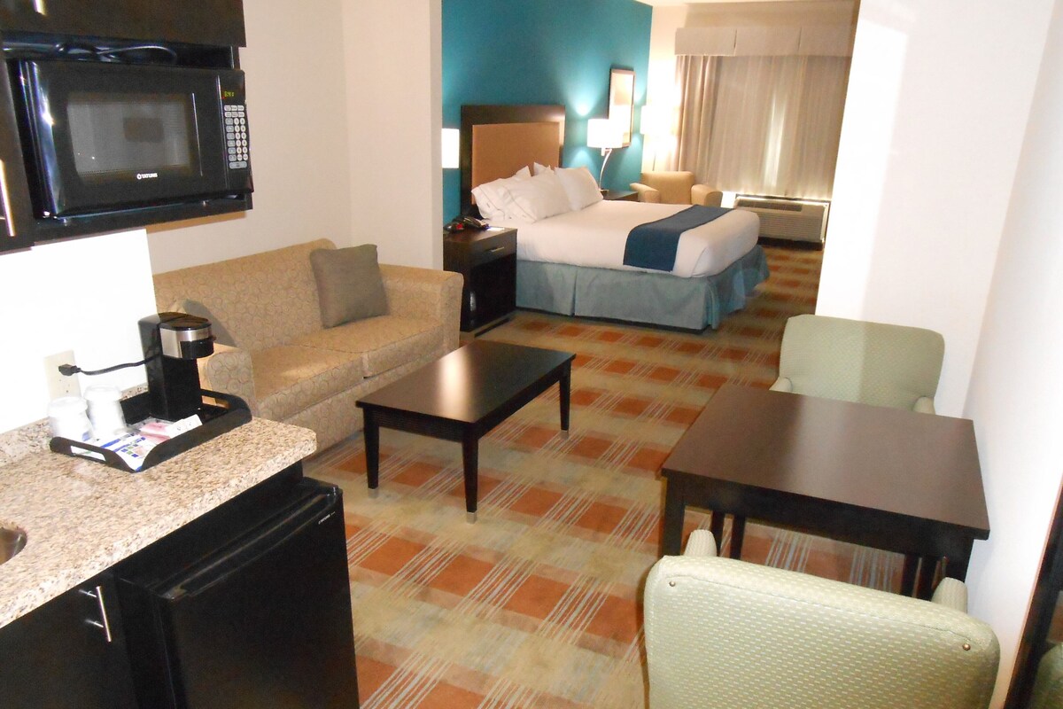 A hotel room with a kitchenette, a bed, a sofa, a dining table, and a chair.