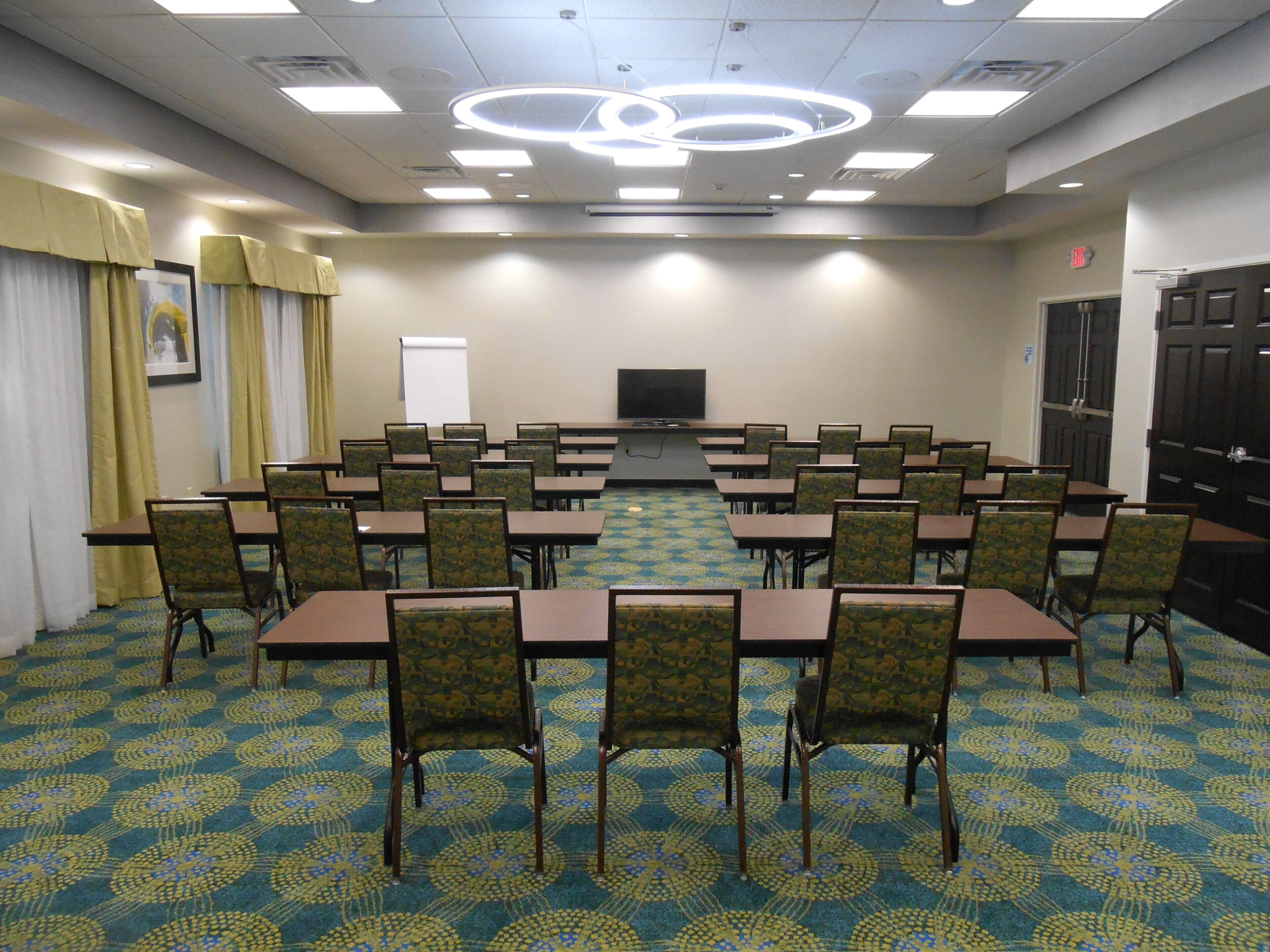 A conference room with tables and chairs arranged in a U-shape, a projector screen, and a patterned carpet.