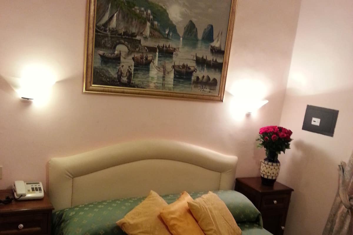 A hotel room with a bed, a nightstand with a telephone, a wall-mounted painting, and a vase with flowers.