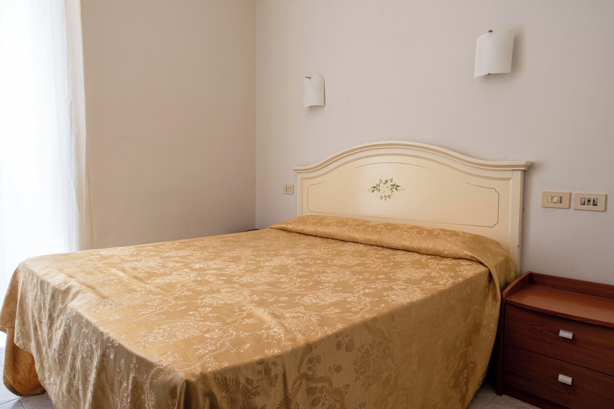 A neatly made bed with a floral bedspread, a wooden nightstand with drawers, and a wall-mounted light fixture.