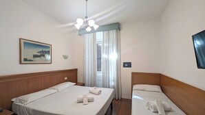 Ein Hotelzimmer mit zwei Betten, einem Holzkopfende, einem Bild an der Wand, einer Deckenleuchte und einem Fenster mit Vorhängen.