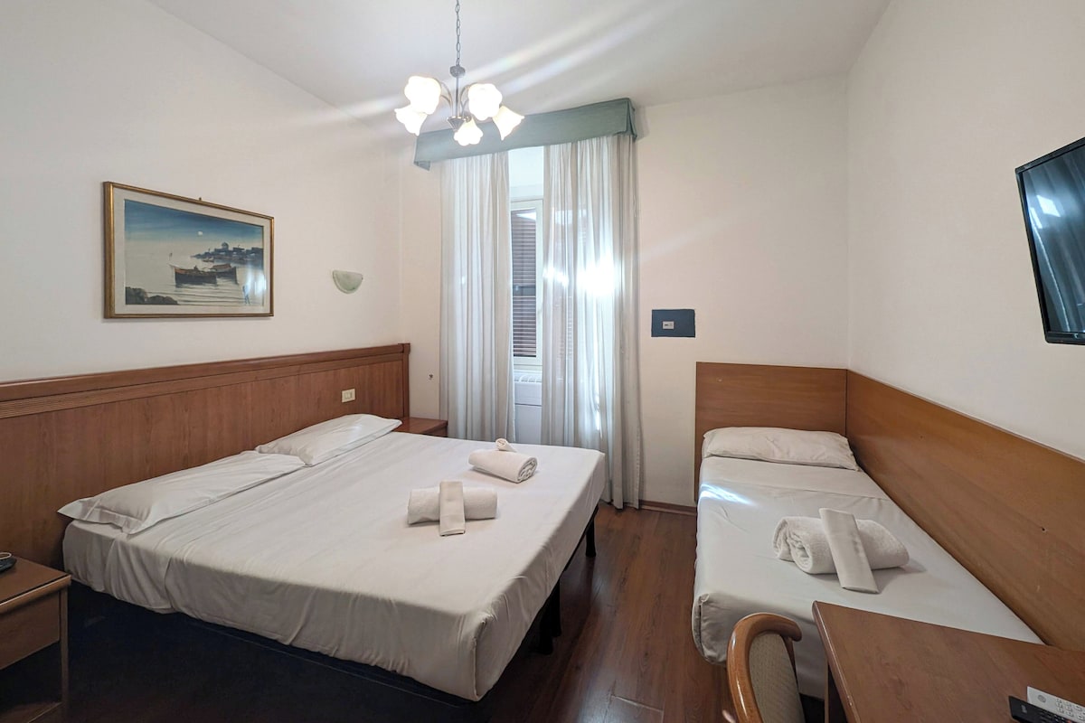 A hotel room with two beds, a wooden headboard, a painting on the wall, a ceiling light fixture, and a window with curtains.