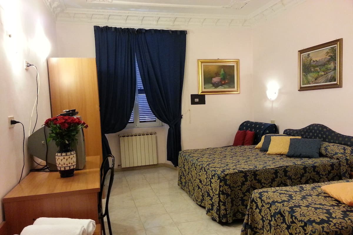 A room with two beds, a desk, a chair, a radiator, a window with curtains, a painting, and a vase with flowers.