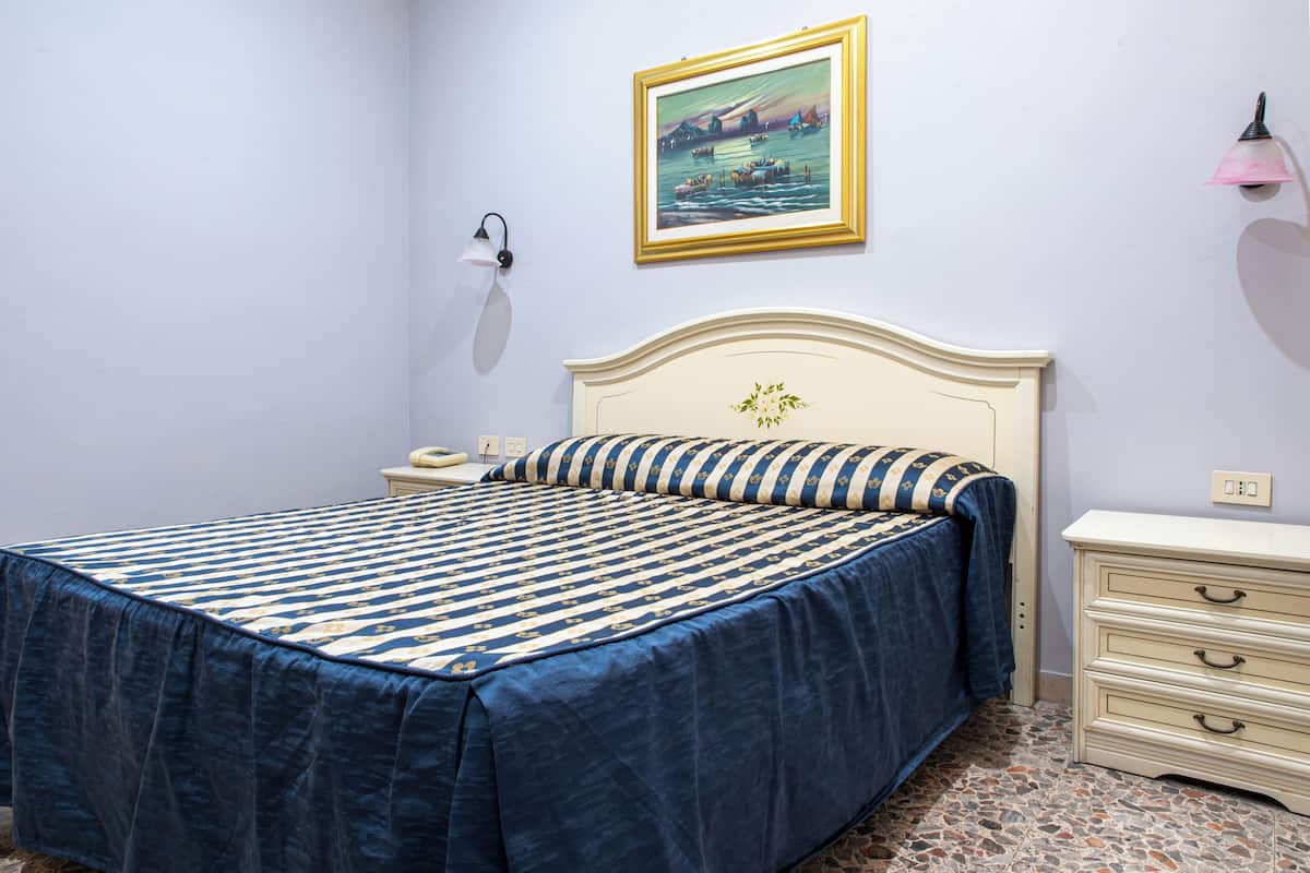 A bedroom with a bed, a nightstand, a dresser, a wall-mounted lamp, and a framed painting.