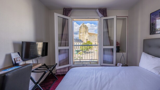 A hotel room with a bed, a TV, a desk, and a window with a view of a building. - Hotel le Beffroi (Dreux)