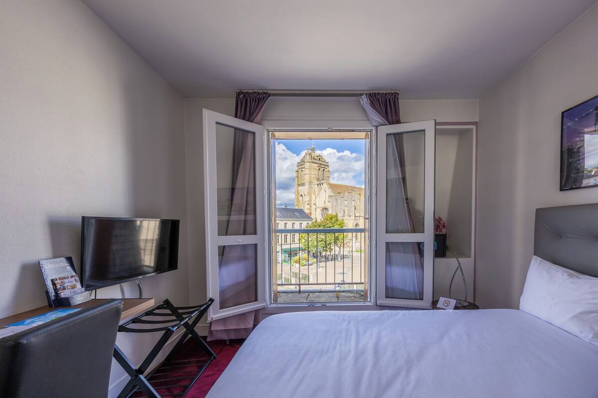A hotel room with a bed, a TV, a desk, and a window with a view of a building.