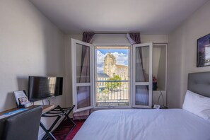 A hotel room with a bed, a TV, a desk, and a window with a view of a building. - Hotel le Beffroi (Dreux)