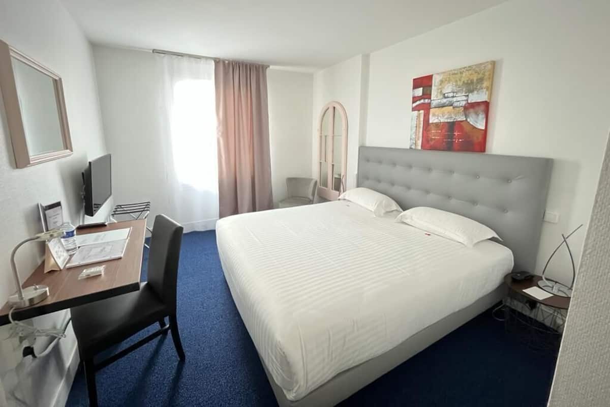 A hotel room with a bed, a desk with a chair, a television, and a window with curtains.