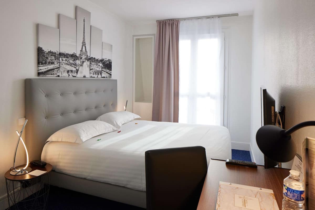 A hotel room with a bed, a desk, a chair, a lamp, a bottle, a remote control, and a window with curtains.