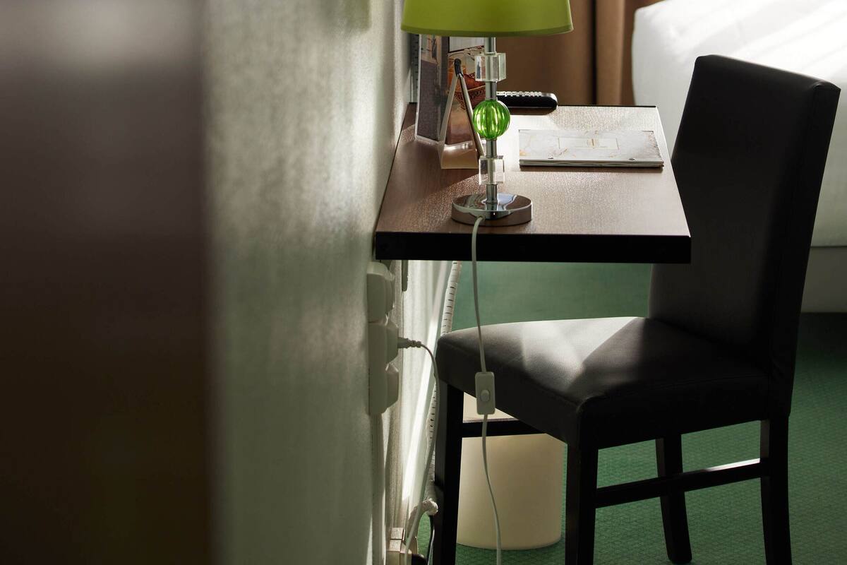 A hotel room with a bed, a desk with a lamp, a chair, and a window with curtains.