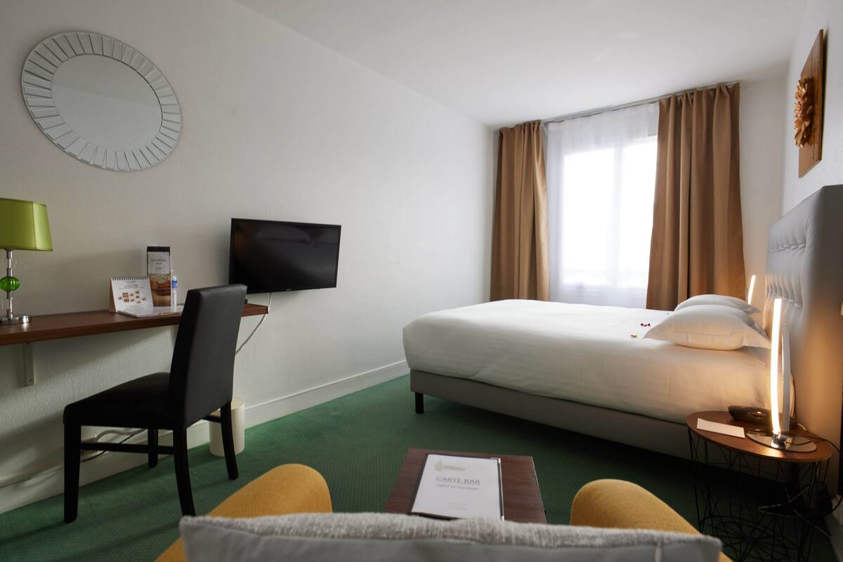 A hotel room with a bed, a desk with a chair, a television, a bedside table with a lamp, and a green carpet.