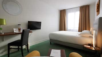 Et hotelværelse med en seng, et skrivebord med stol, et fjernsyn, et sengebord med lampe og et grønt tæppe.
