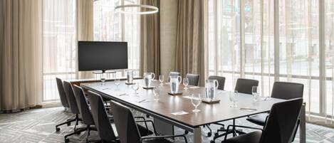 Un salón de conferencias con una larga mesa, sillas, un televisor de pantalla plana y ventanas grandes con cortinas.