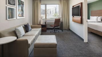 A hotel room with a sofa, ottoman, desk, chair, TV, and a bed.