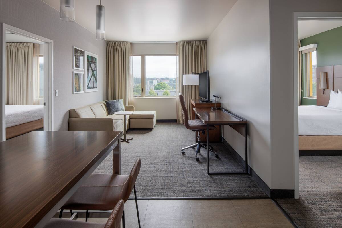Una habitación de hotel con cama, escritorio con silla, sofá, televisor y ventana con cortinas.