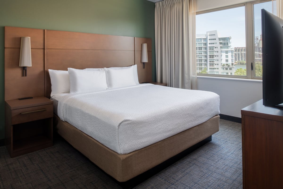 Habitación de hotel con una cama grande, una mesita de noche, un televisor sobre un mueble y una ventana con vistas a edificios.