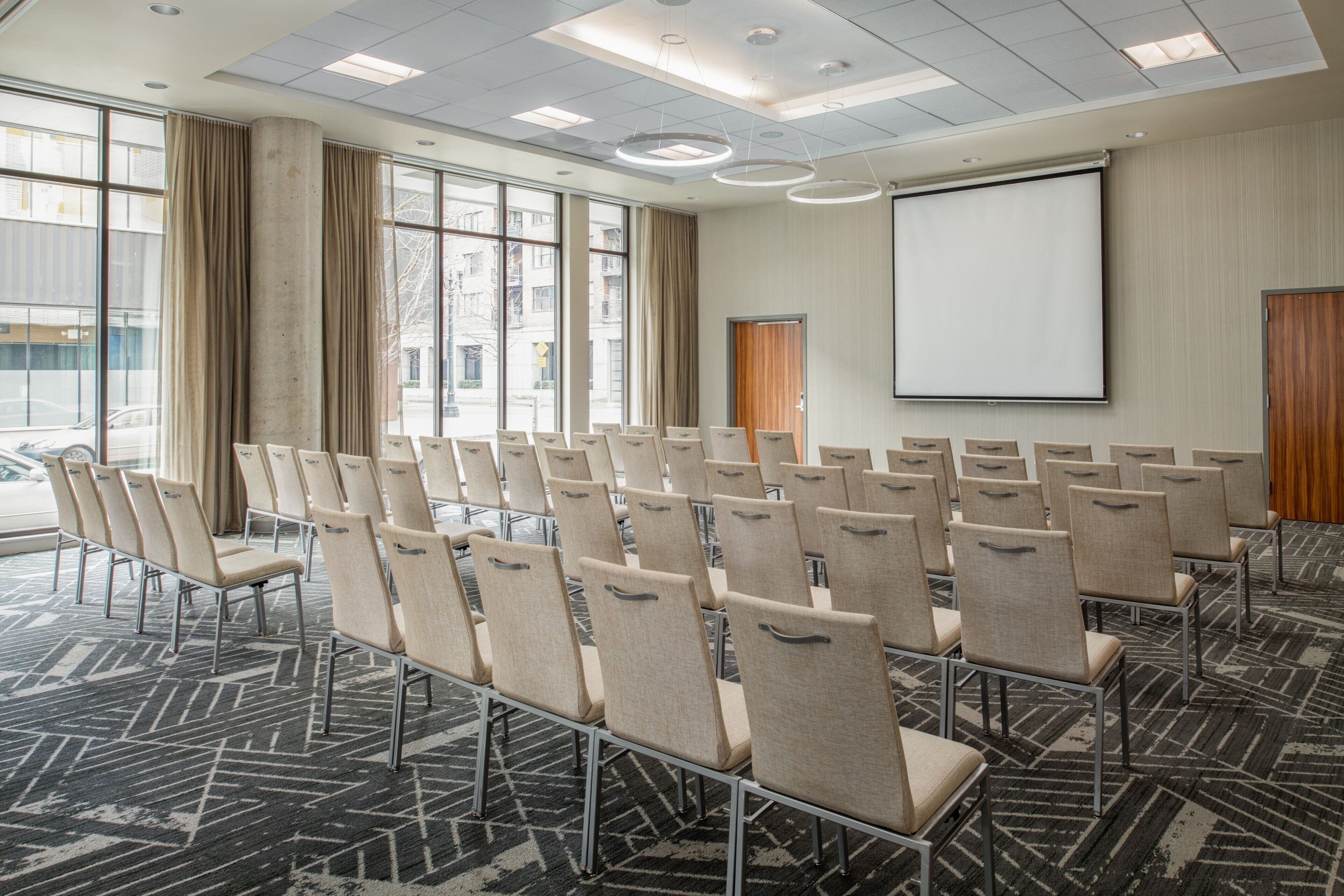 Une salle de conférence avec des rangées de chaises, un écran de projection et de grandes fenêtres.