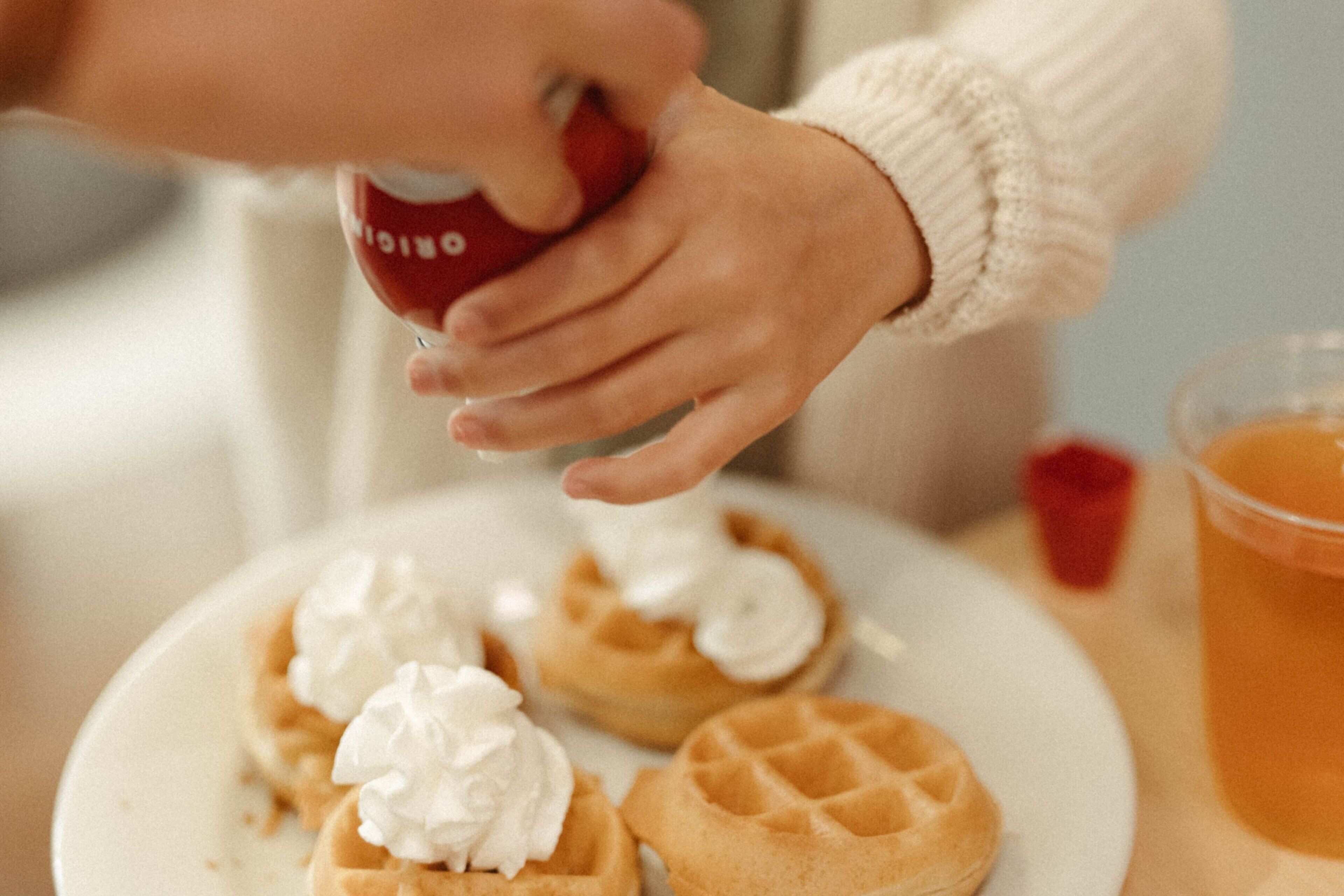 A close-up of a hand pouring whipped cream onto a waffle.
