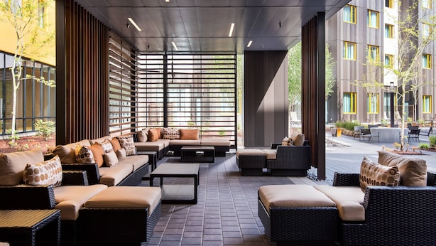 Un espace de détente extérieur moderne, avec des meubles en osier, un sol carrelé et de grandes fenêtres offrant une vue sur les bâtiments environnants.