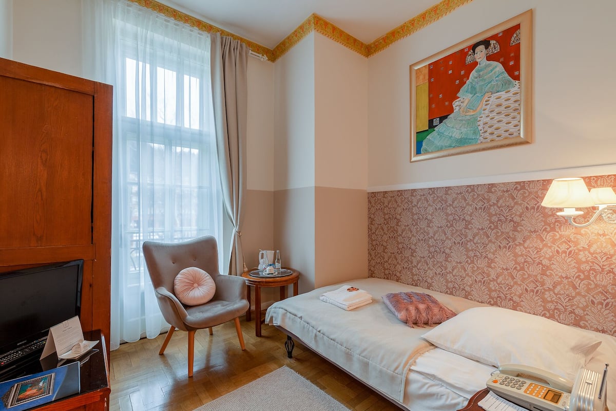 A hotel room with a bed, a chair, a TV, a table with a lamp, and a painting on the wall.