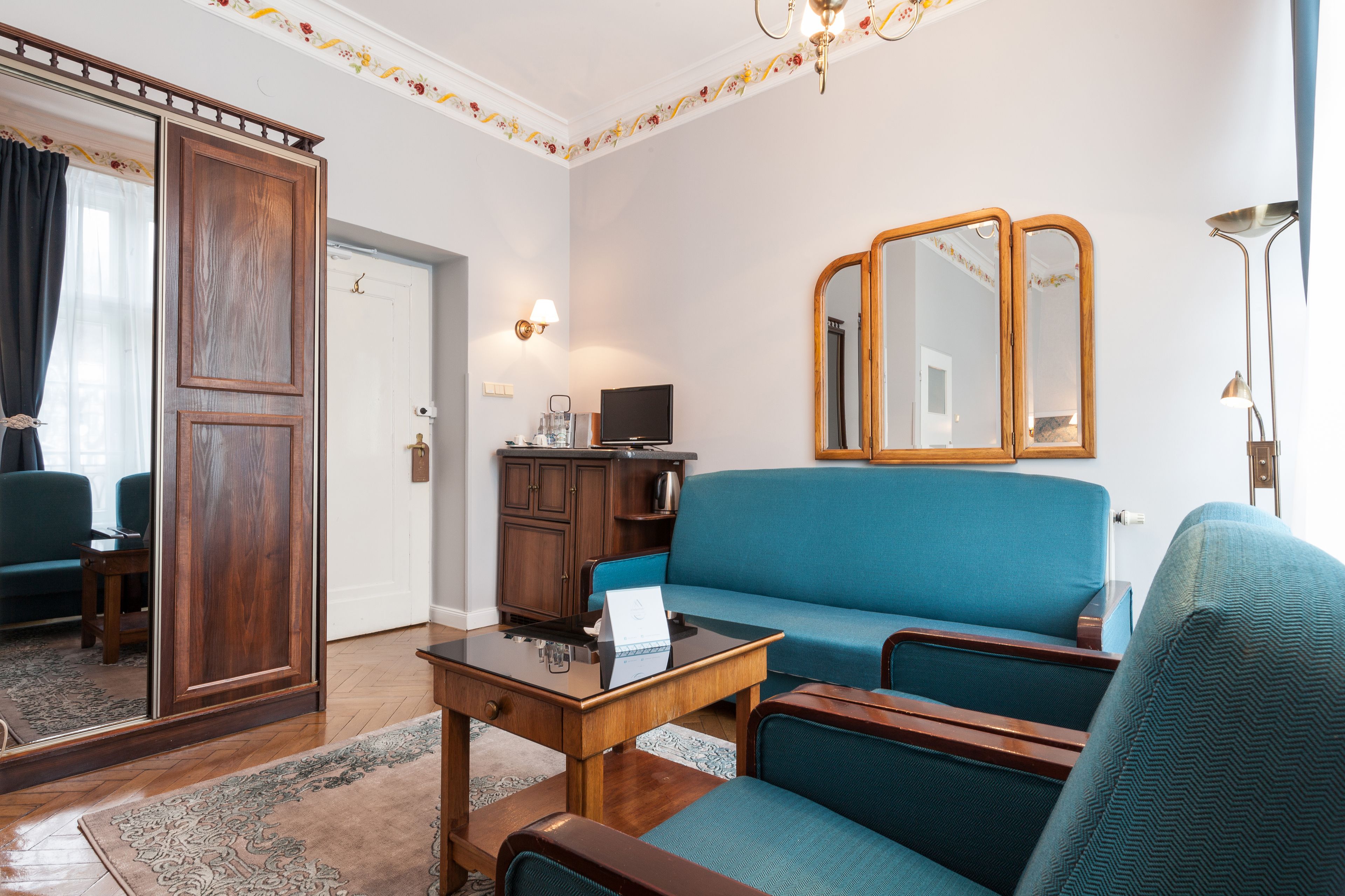 Una sala de estar con un sofá azul, una mesa de centro de madera y un televisor. Se ve un armario con espejo, una lámpara y una alfombra en el suelo.