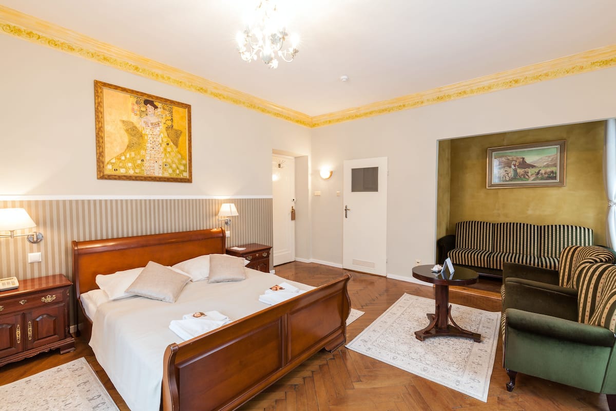 A hotel room with a large bed, a green armchair, a painting on the wall, and a chandelier.