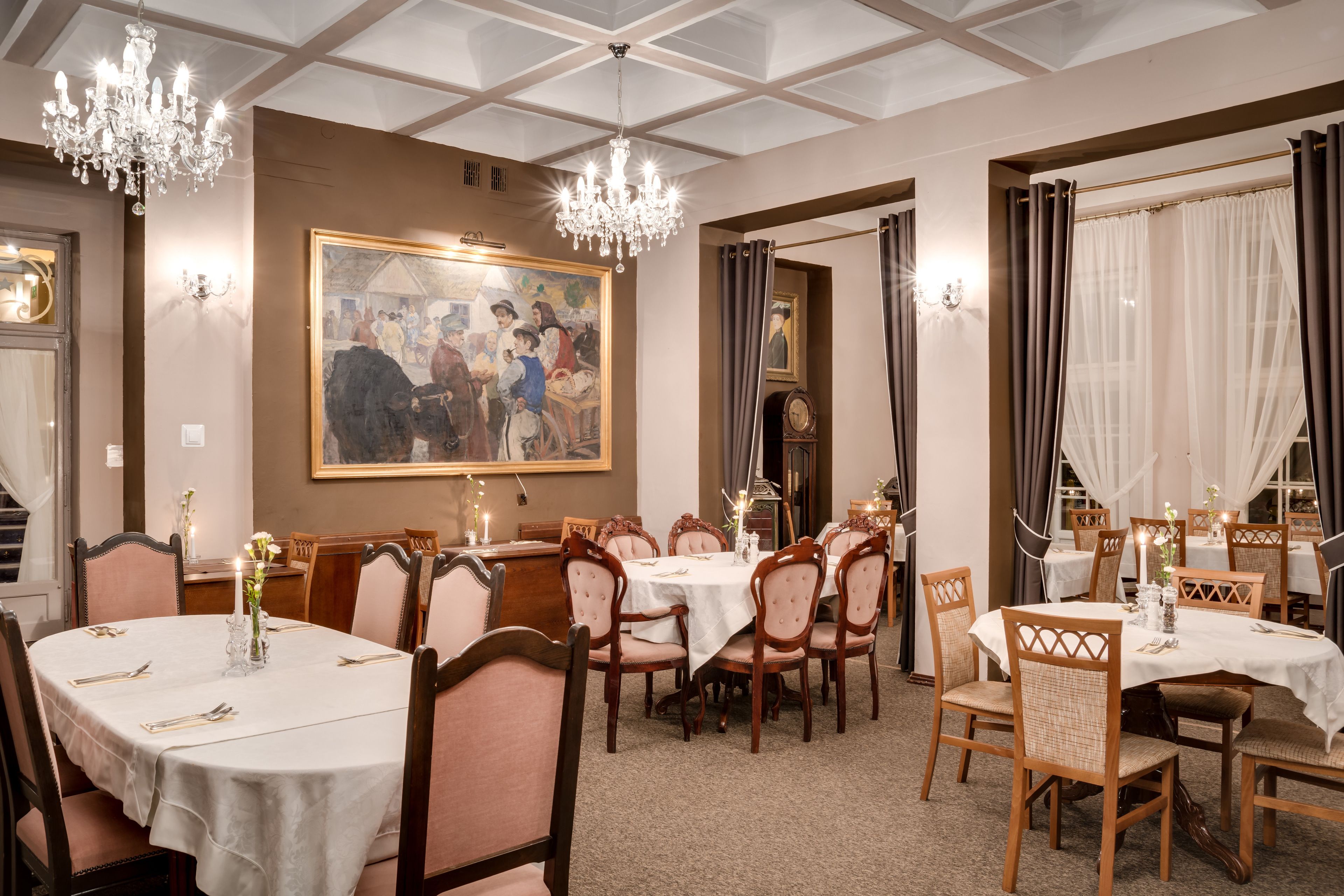 A dining area with round tables, wooden chairs, chandeliers, and a large painting on the wall.