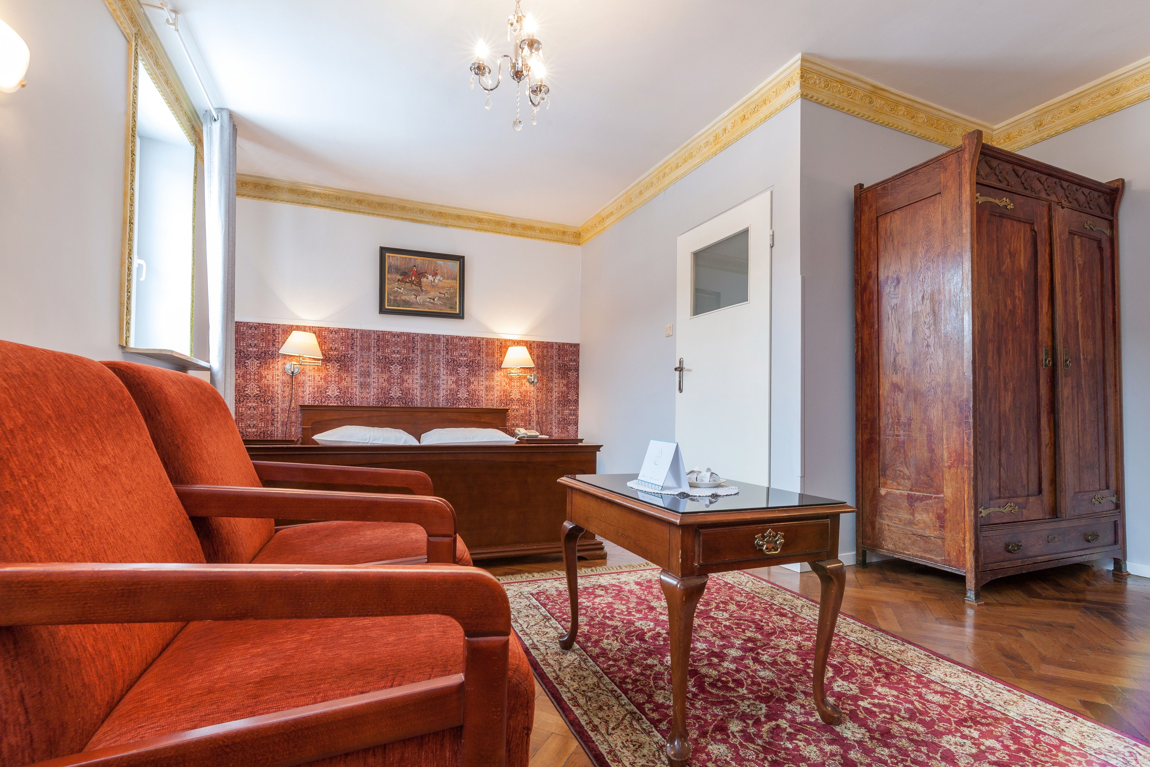 Une chambre d’hôtel avec une grande armoire en bois, un lit avec une tête de lit rouge, deux lampes de chevet et un fauteuil rouge.