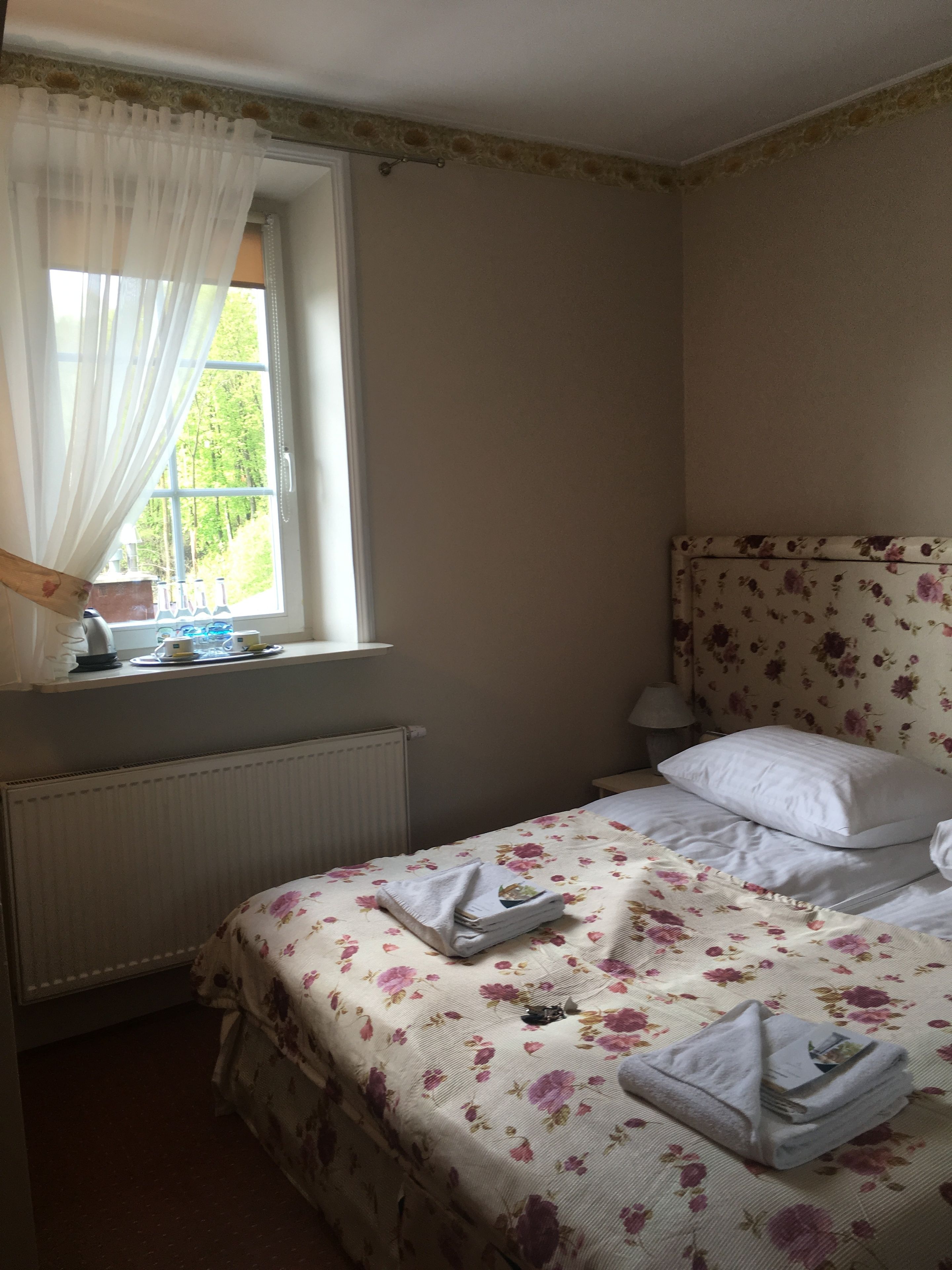 Une chambre à coucher avec une couvre-lit à motifs floraux, un lit bien fait avec des oreillers blancs, une fenêtre avec des rideaux légers, une table de chevet avec une lampe et un radiateur.