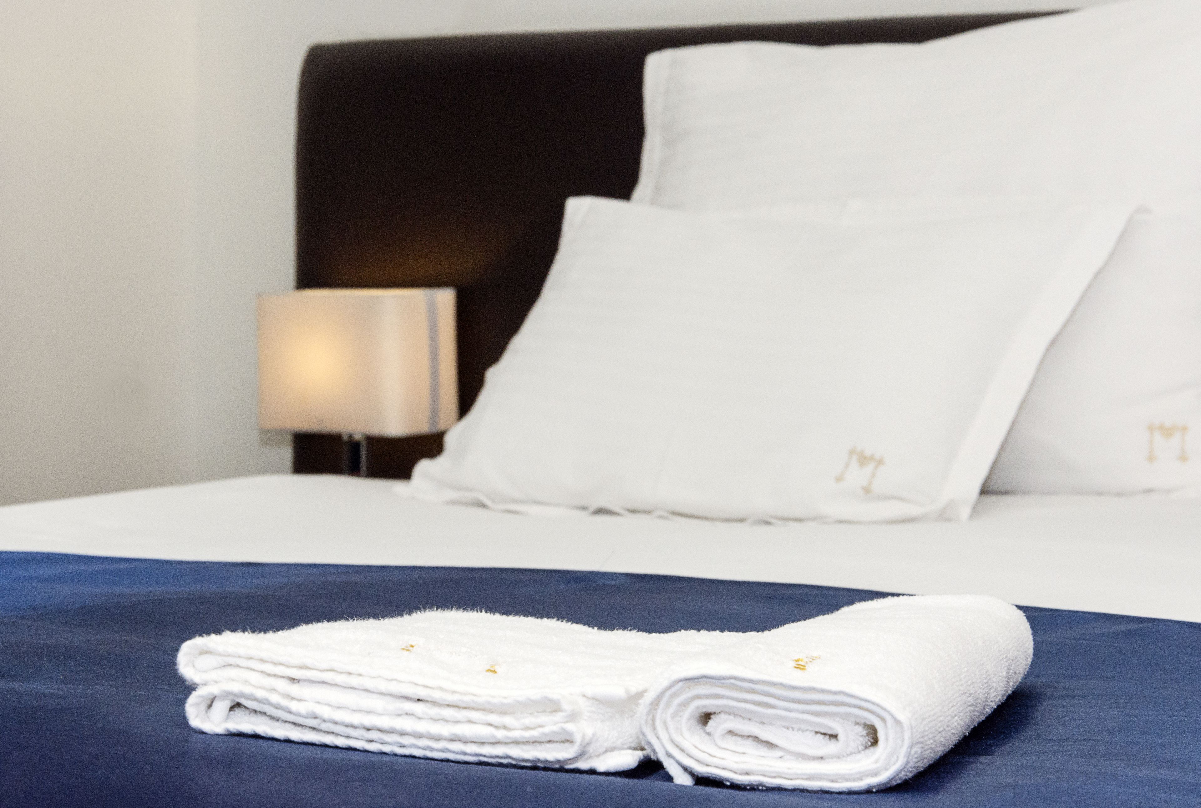 Una cama bien hecha con toallas blancas y una colcha azul, una almohada blanca y una lámpara de noche.