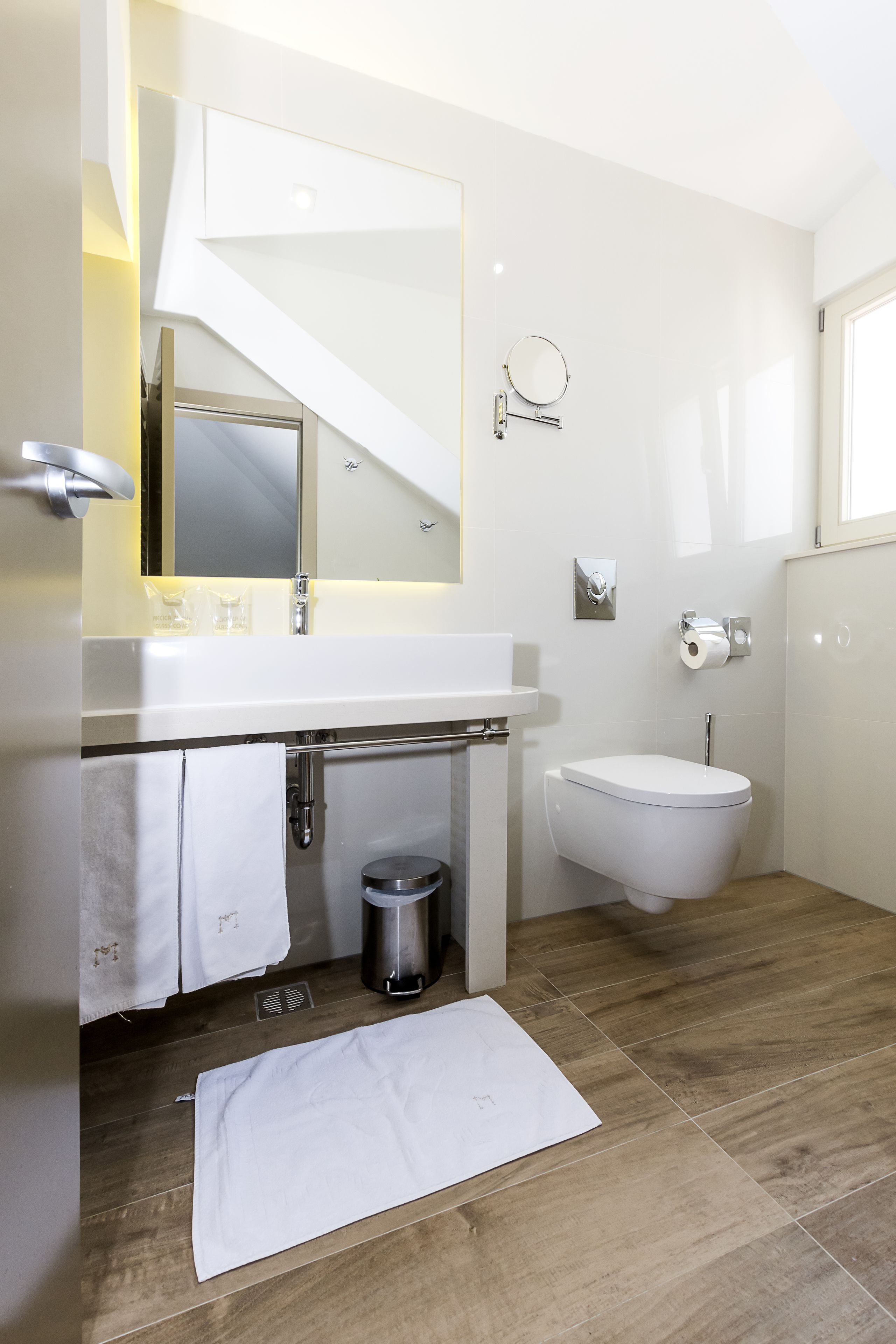 A modern bathroom with a white sink, mirror, and toilet. There is a towel rack with towels, a trash bin, and a small mat on the floor.